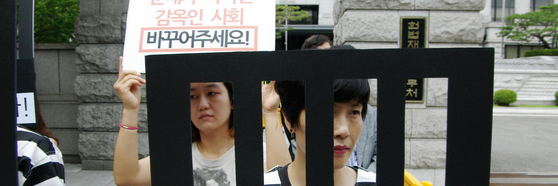 Eteläkorealainen aseistakieltäytyjä pitää kiinni pahvistista tekokaltereista ja on pukeutunut musta-valko-raidallisiin vanginvaatteisiin.