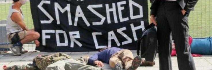 Maassa makaa levällään ihmisiä, jotka esittävät kuolleita. Etualalla pukuun pukeutunut mies katselee kohti kameraa silmälasiensa yli. Takana aktivistit pitävät banderollia: Human lives smashed for cash. 