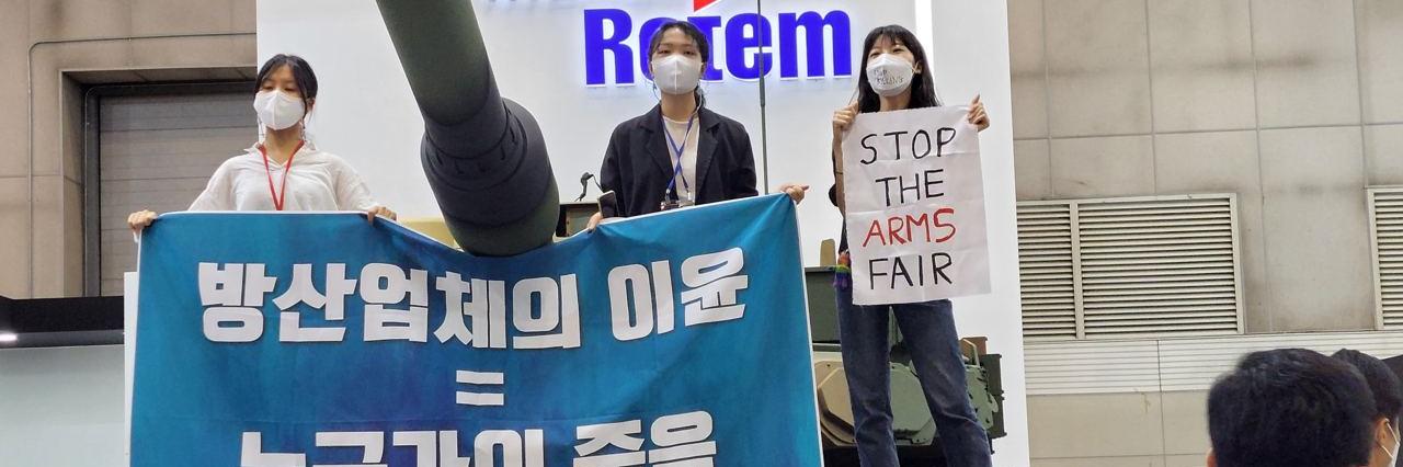 Three activists step up on a tank holding a banner that reads Profits of Arms Industry equals Death of Someone, in an arms fair held in Korea