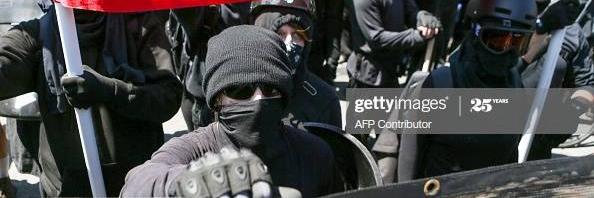 Mustiin pukeutuneet ja huivein, kilvin ja kypärin varustautuneet mielenosoittajat kantavat banderolleja ja punamustaa anarkistilippua.