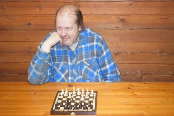 Kaj Raninen istuu pöydän ääressä hirsiseinäisessä huoneessa nojaten käteensä. Hänen edessään on shakkilauta.