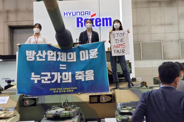 Three activists step up on a tank holding a banner that reads Profits of Arms Industry equals Death of Someone, in an arms fair held in Korea