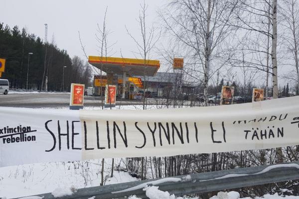 Kuvassa näkyy Shellin huoltoasema Tampereella. Etualalla Elokapinan logoilla varustettu banderolli, jossa lukee: "Shellin synnit ei mahdu tähän"